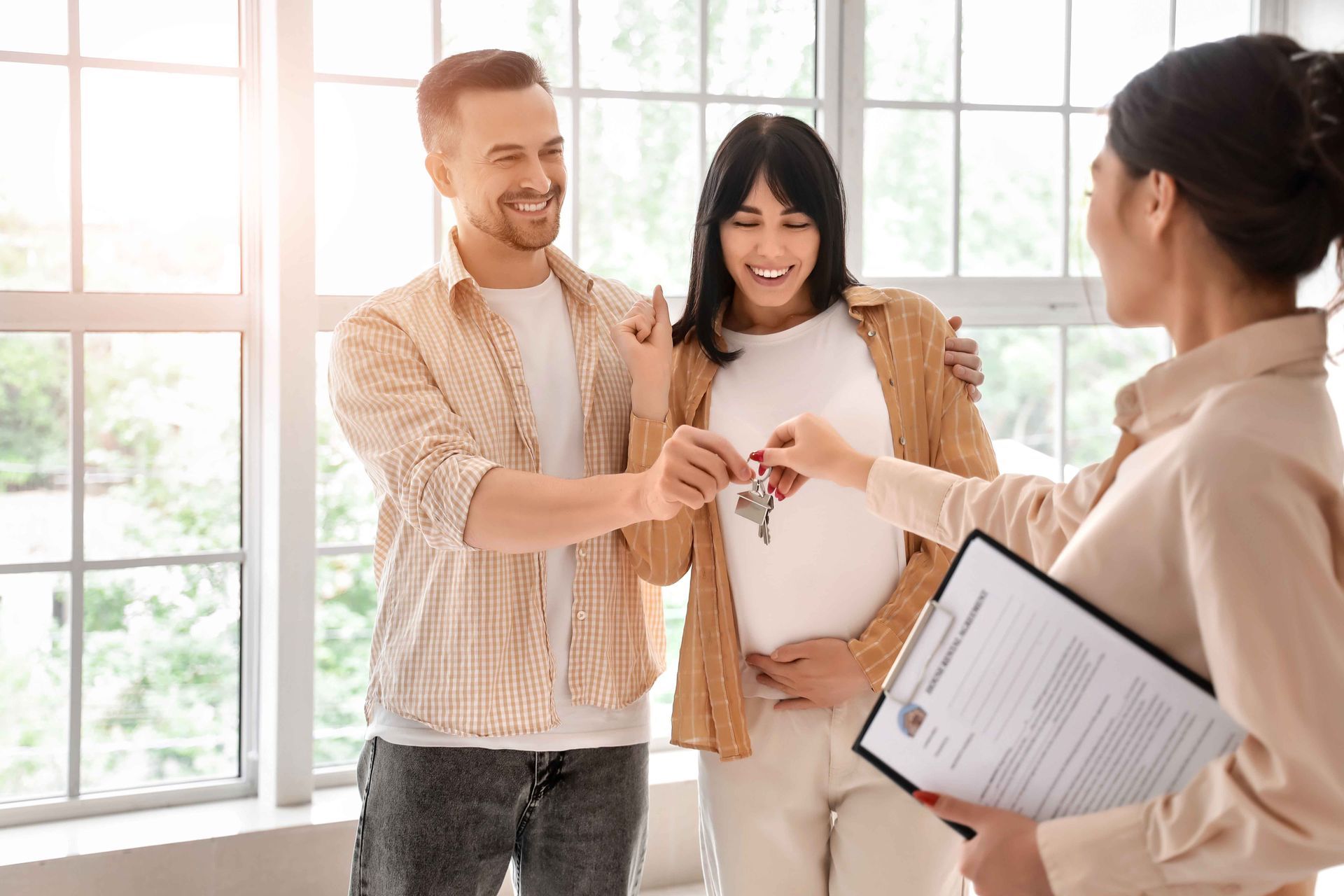 Un agent immobilier remet les clés à un couple souriant dans une pièce lumineuse avec des fenêtres.