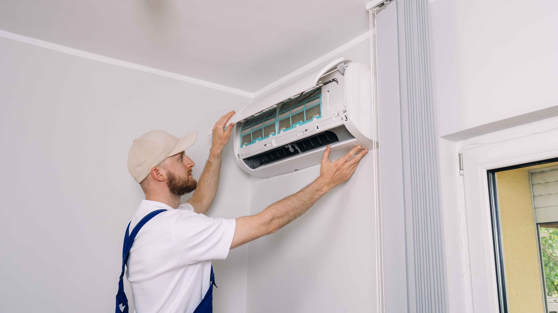 Un technicien installe un climatiseur mural à l'intérieur, vêtu d'une chemise blanche et de bretelles bleues.