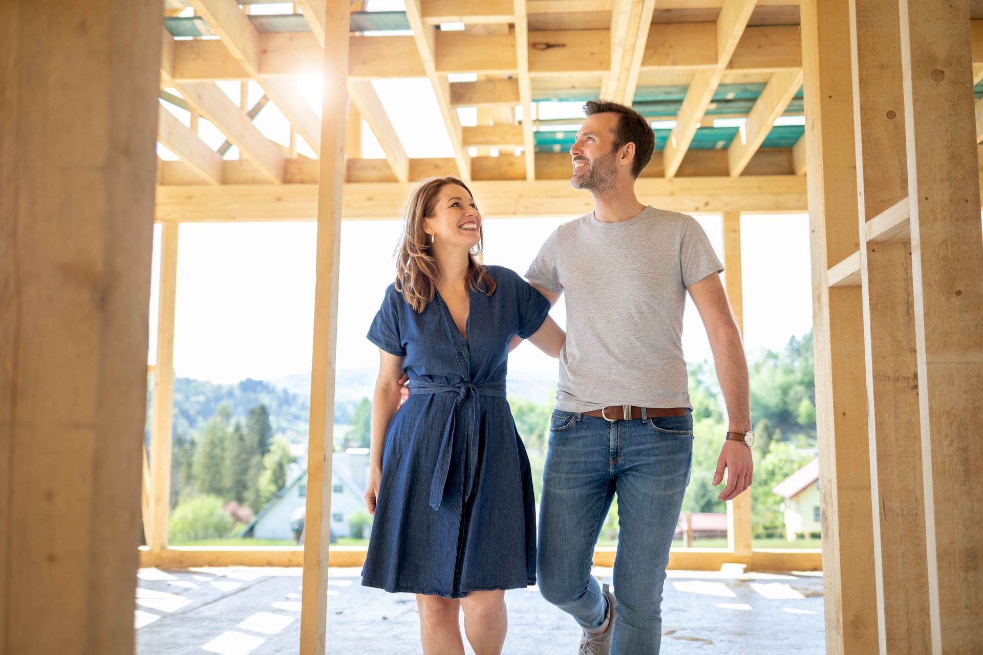 Un couple souriant traverse une maison en construction ; charpente en bois, cadre ensoleillé.