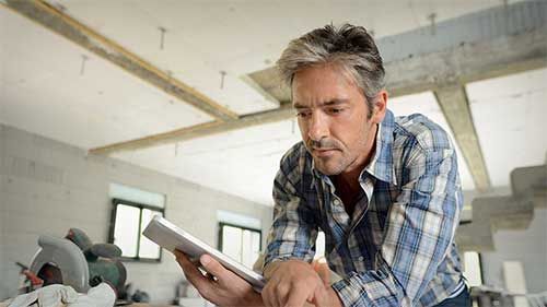 Un homme en chemise à carreaux regarde une tablette dans une pièce en construction.