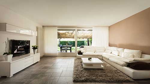 Salon moderne avec canapé d'angle blanc, télévision et grande fenêtre donnant sur une terrasse.