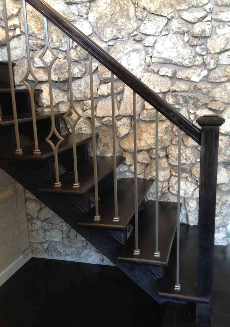 Dark wooden staircase with black railing against a rough stone wall.