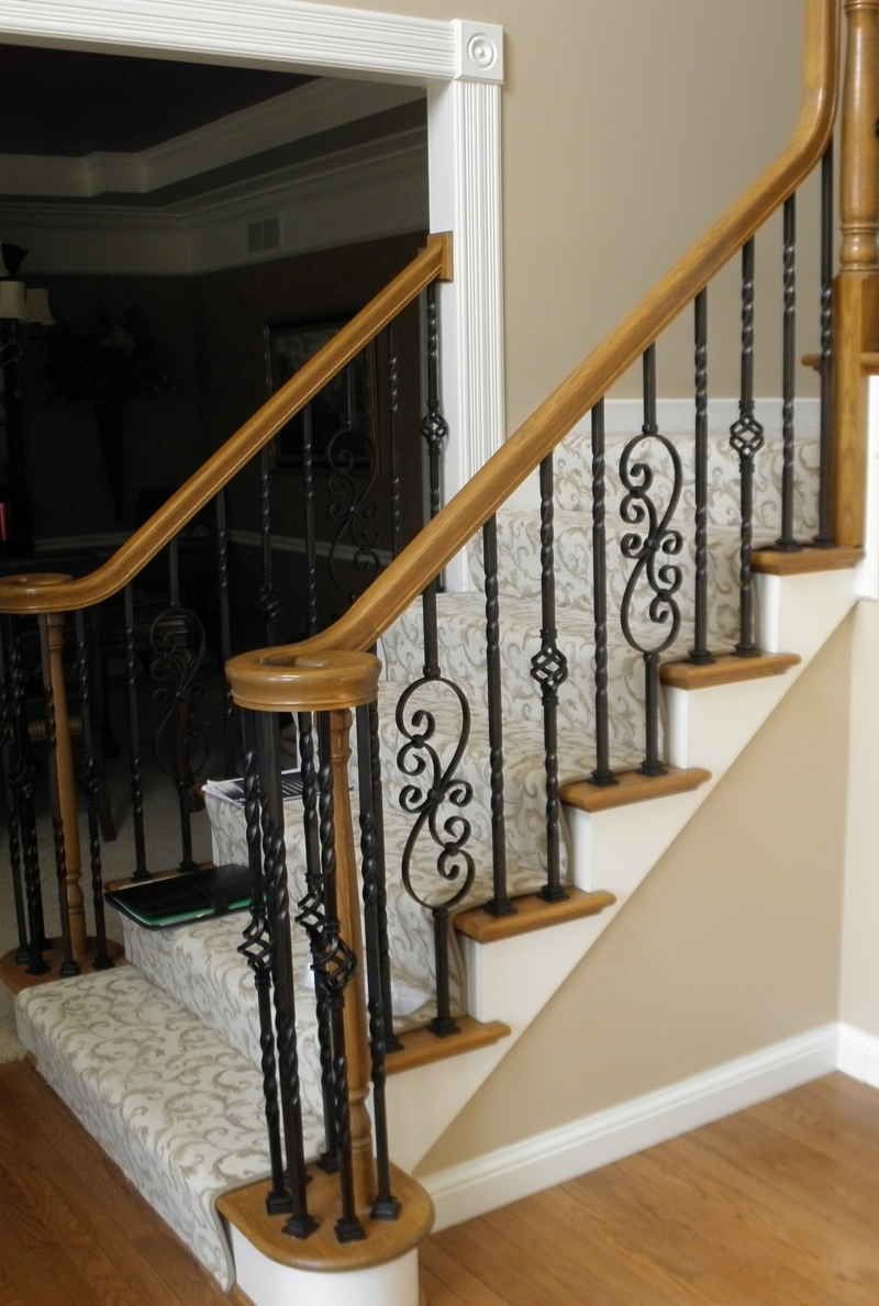 Staircase with wood railing, black wrought-iron balusters, and beige carpeted steps beside a hardwood floor.