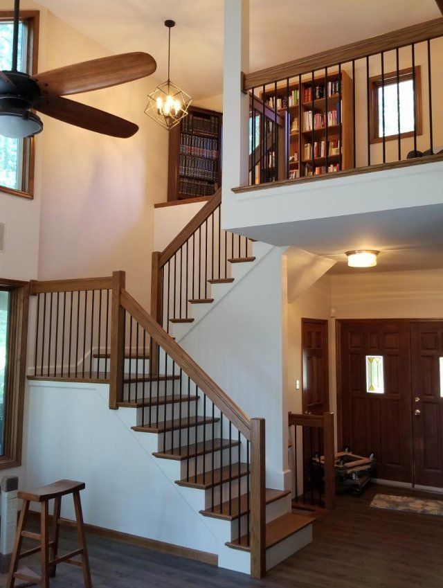 Interior staircase with white walls, wooden railings, loft bookshelf, and ceiling fan in a bright home foyer