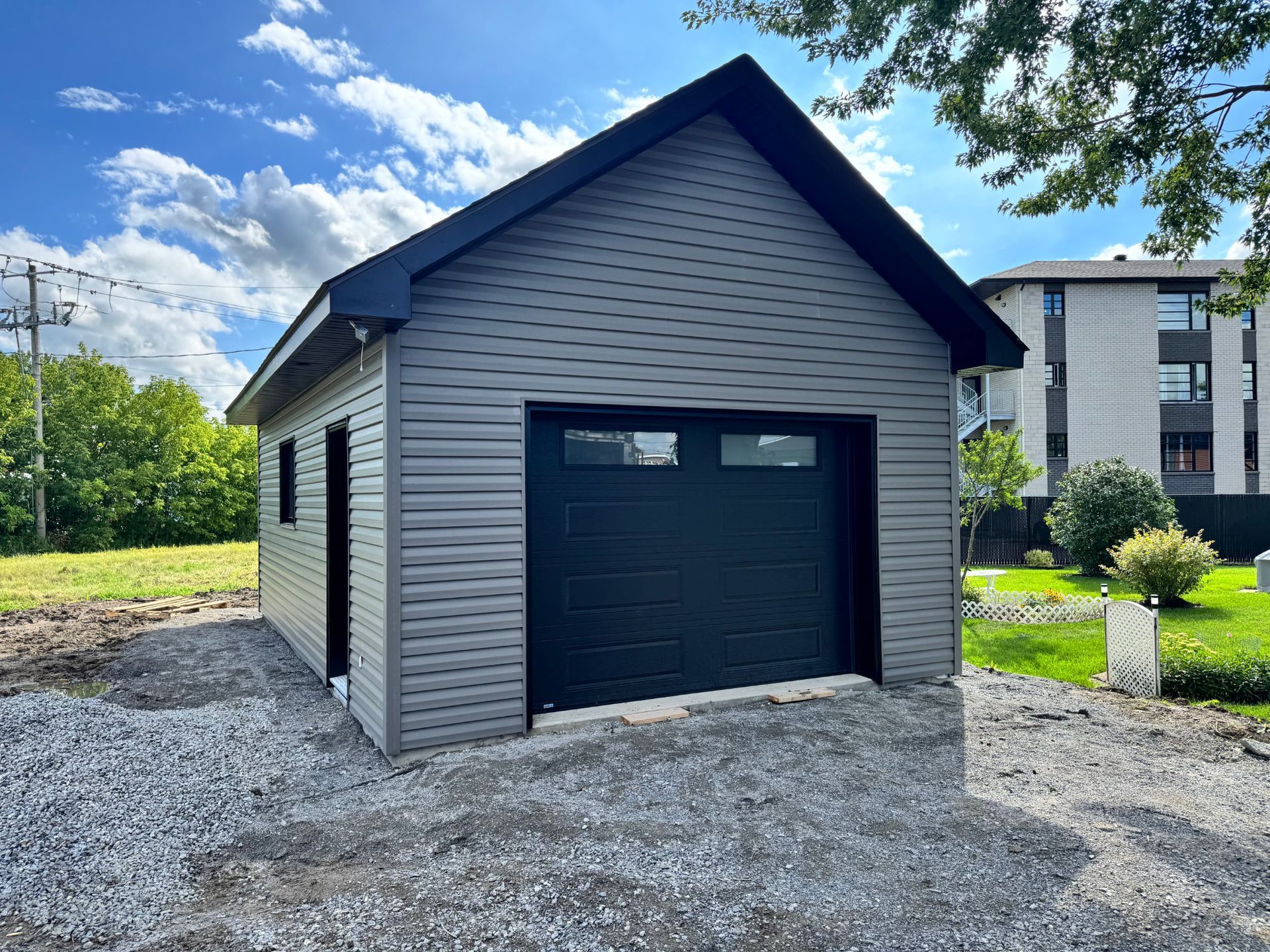 Petit garage gris indépendant avec une porte noire, sur un terrain gravillonné sous un ciel bleu.