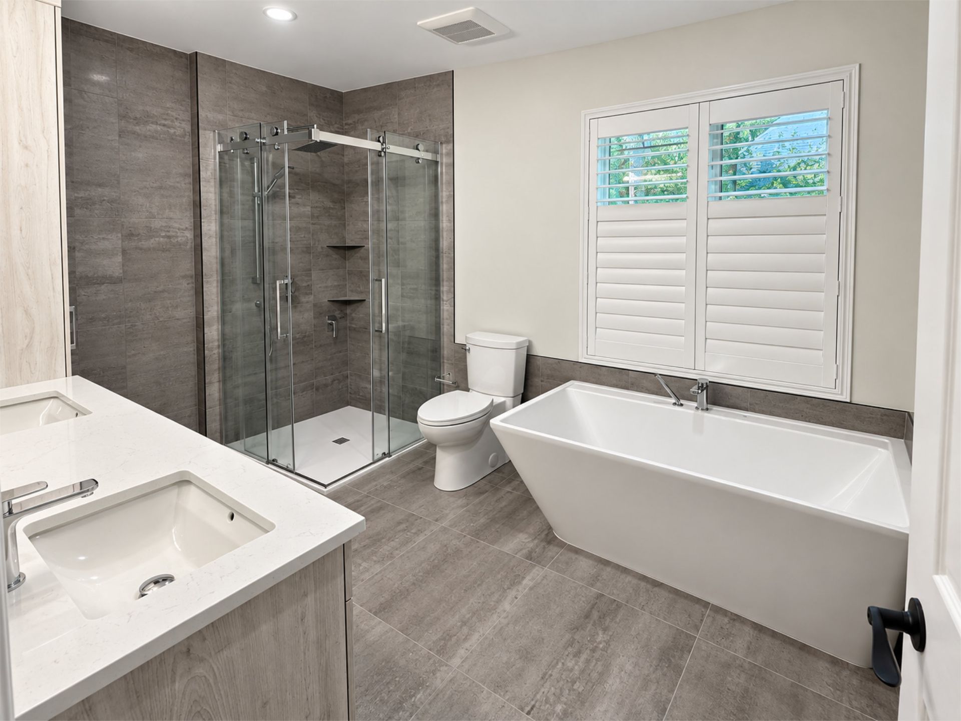 Salle de bains moderne avec double vasque, douche vitrée, baignoire îlot et fenêtre à volets laissant entrer la lumière naturelle