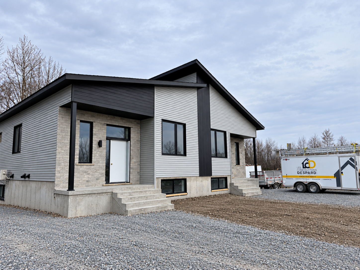 Maison moderne blanche en construction, avec une allée en gravier et une remorque utilitaire à proximité