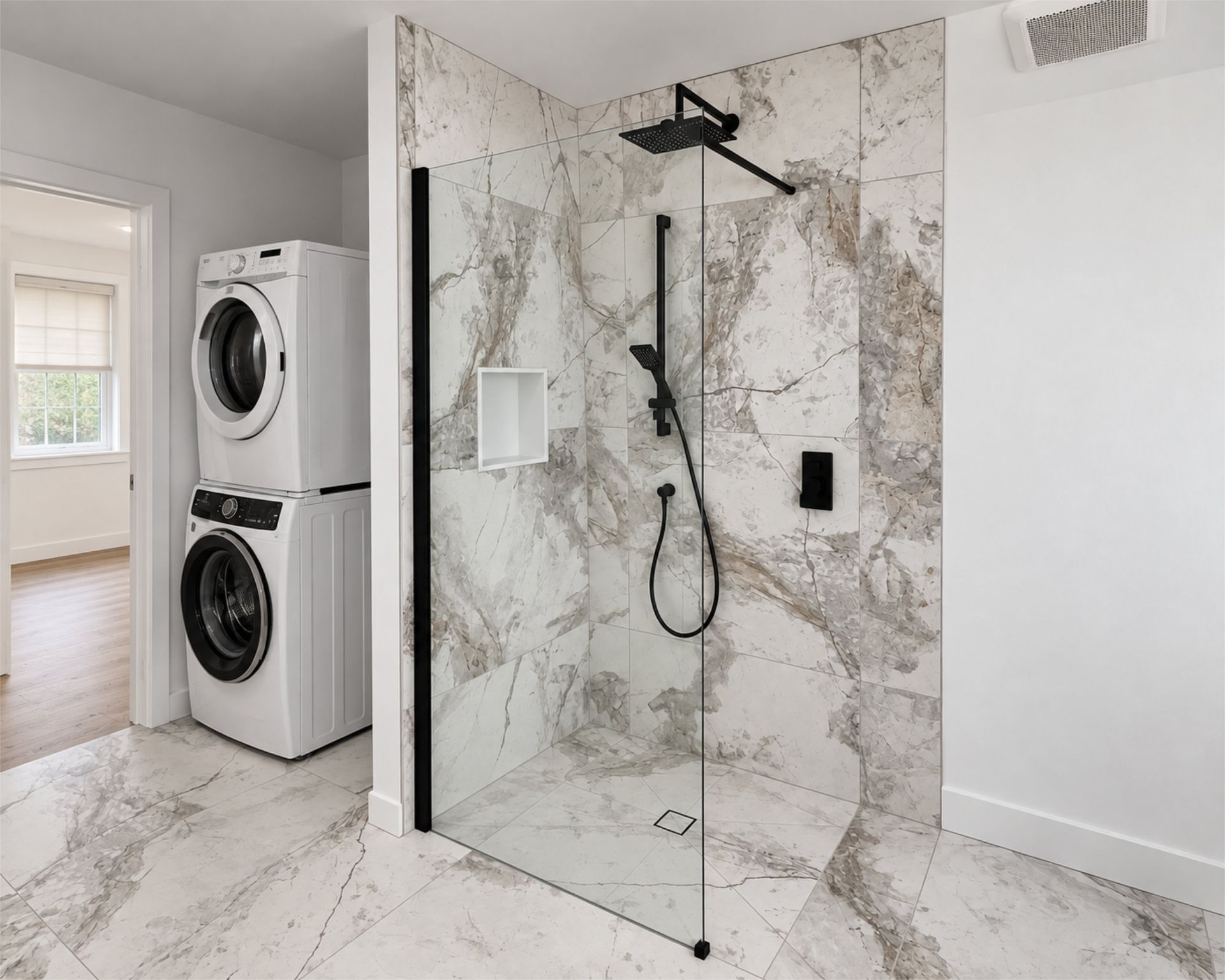 Salle de bains moderne avec douche en marbre et robinetterie noire, à côté d’un lave-linge et d’un sèche-linge superposés