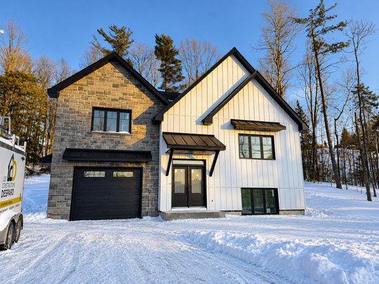 Maison moderne à deux étages, avec un garage noir et un bardage blanc, dans un cadre boisé enneigé