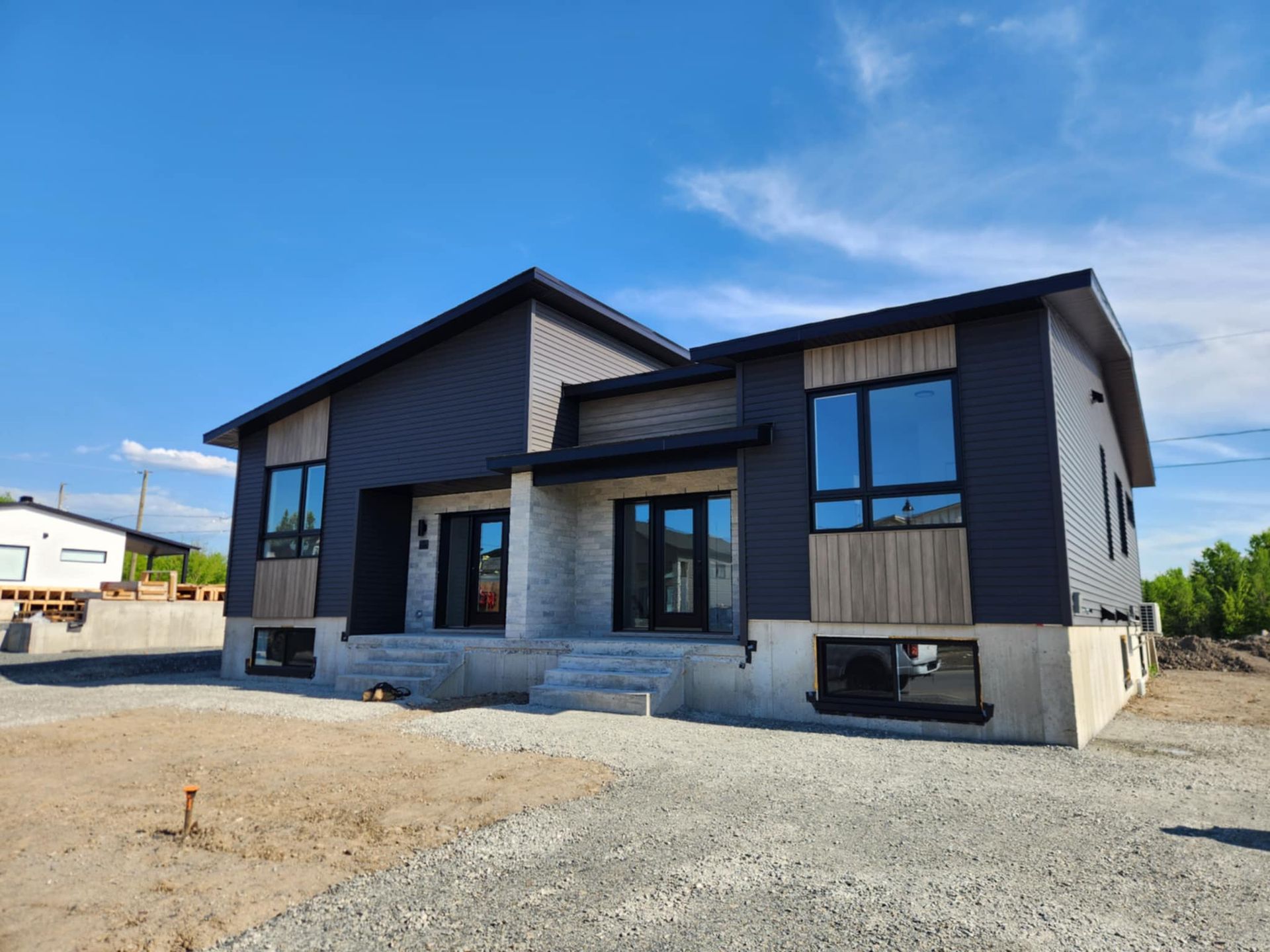 Façade moderne d’une maison grise avec de grandes fenêtres et un porche à l’avant, sous un ciel bleu