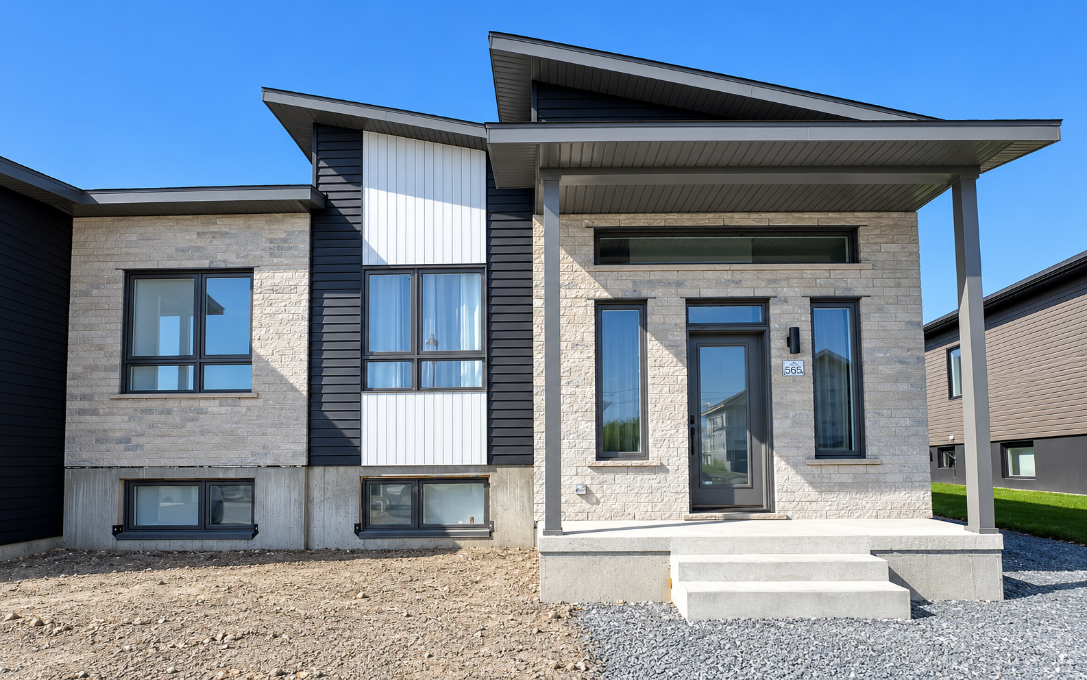 Maison moderne en briques grises avec un porche couvert à l’avant et de grandes fenêtres sous un ciel bleu