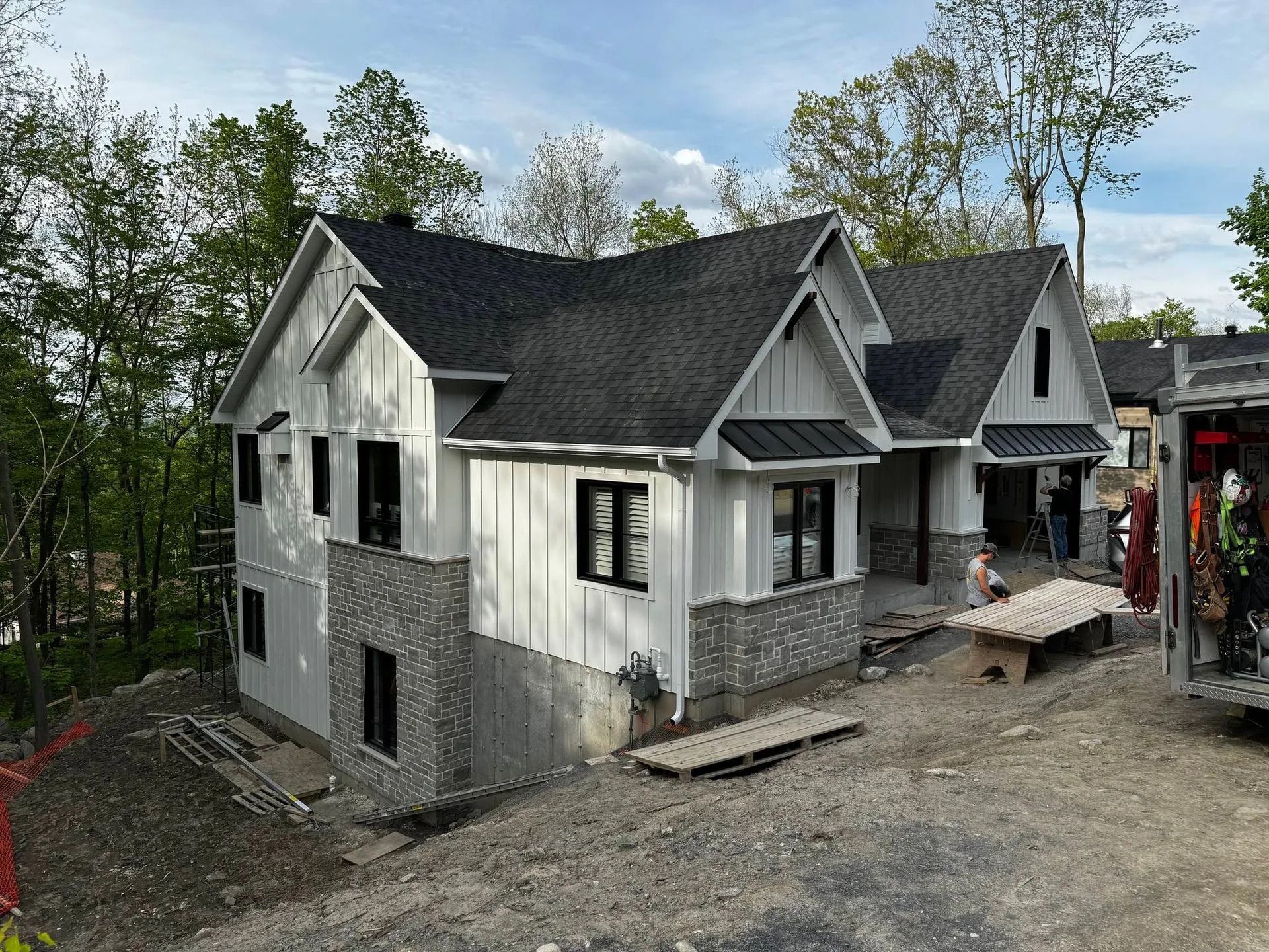 Maison blanche en construction, avec un toit gris et un soubassement en pierre, entourée de terre et d’arbres