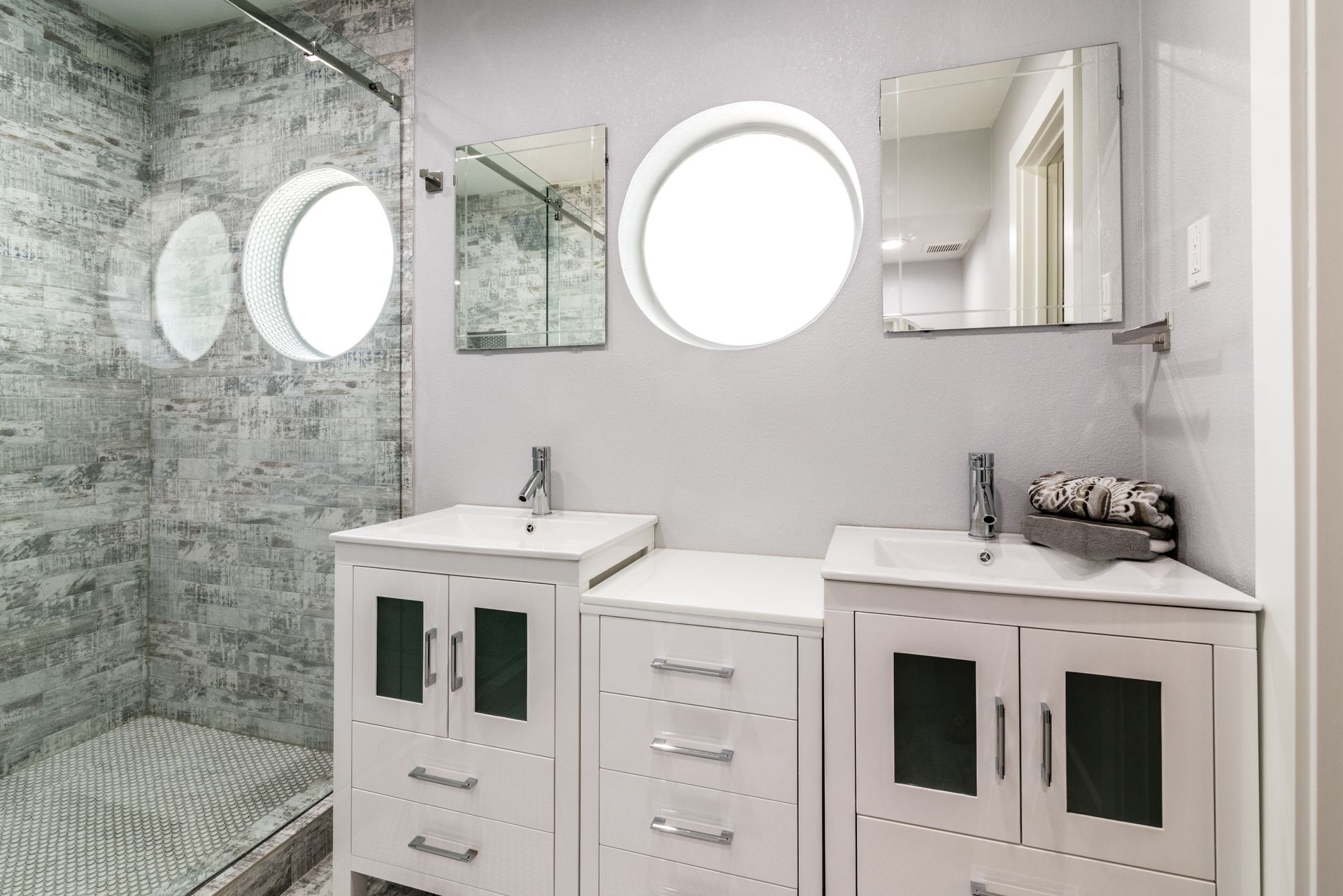 Bright bathroom with glass shower, white vanity cabinets, round mirrors, and gray stone tile.