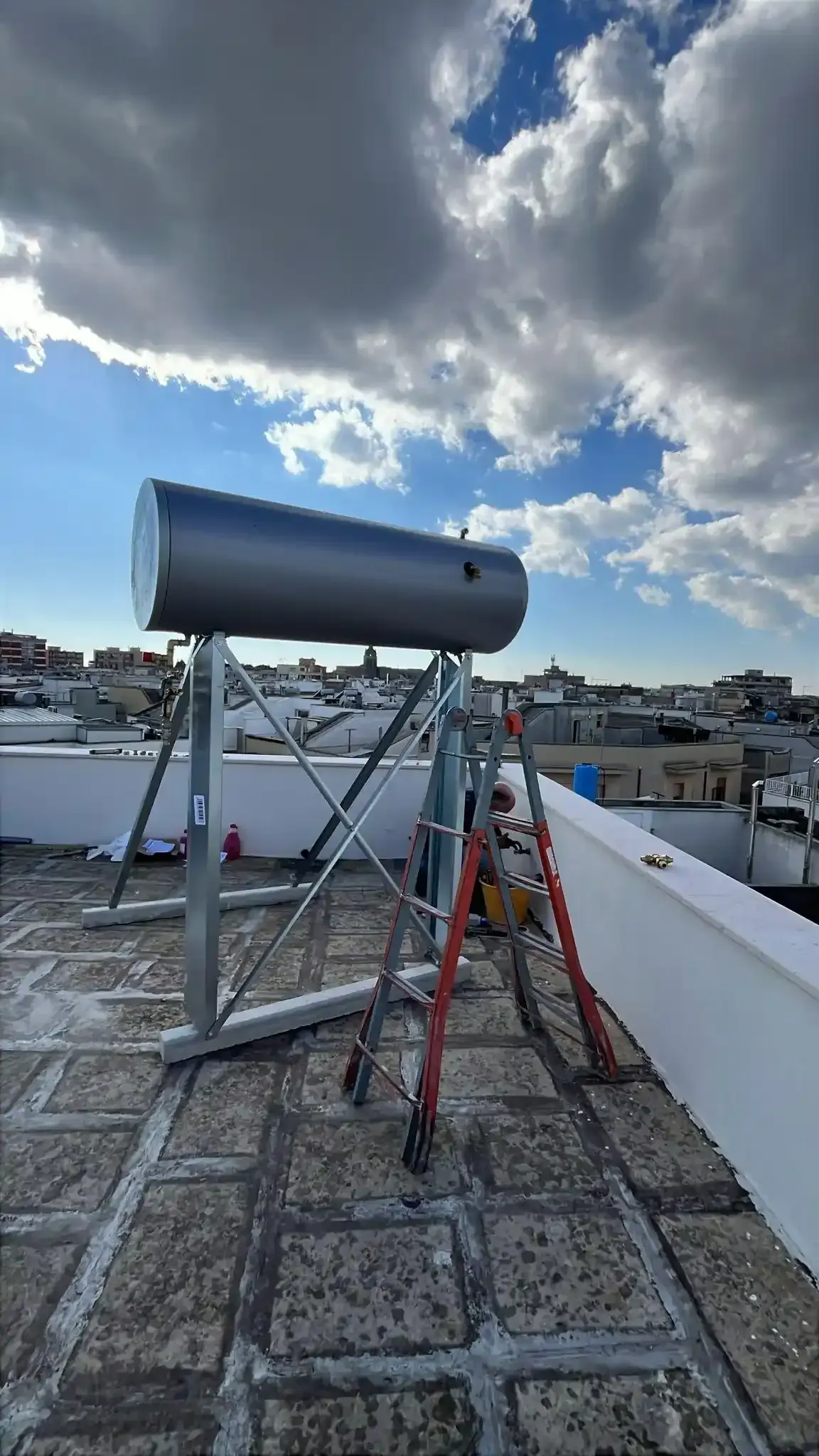 Serbatoio per scaldabagno solare installato sul tetto, su una struttura metallica, sotto un cielo nuvoloso, con gli edifici della città sullo sfondo.
