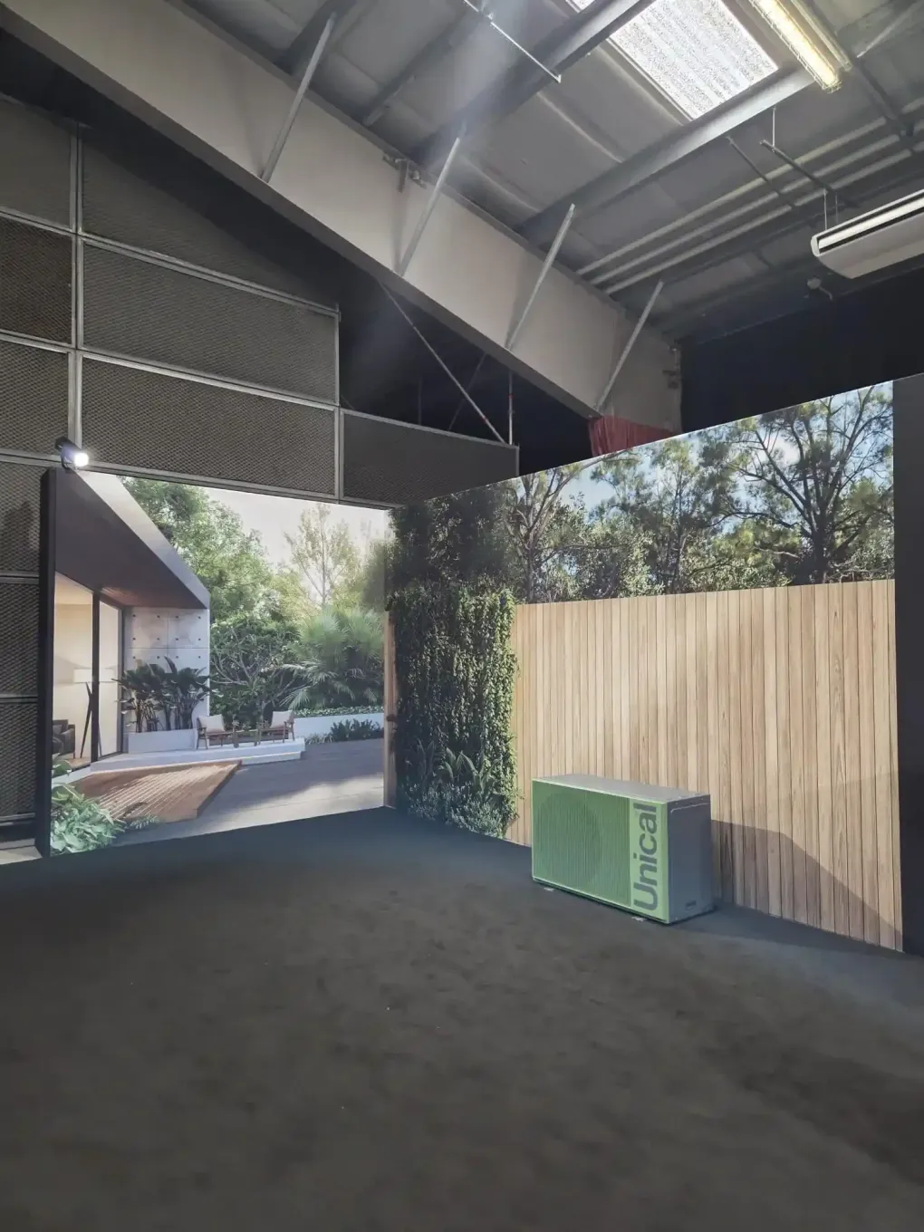 Spazio eventi al coperto con grandi grafiche murali a tema giardino, uno sfondo con una recinzione in legno e una cassa verde su un pavimento scuro.