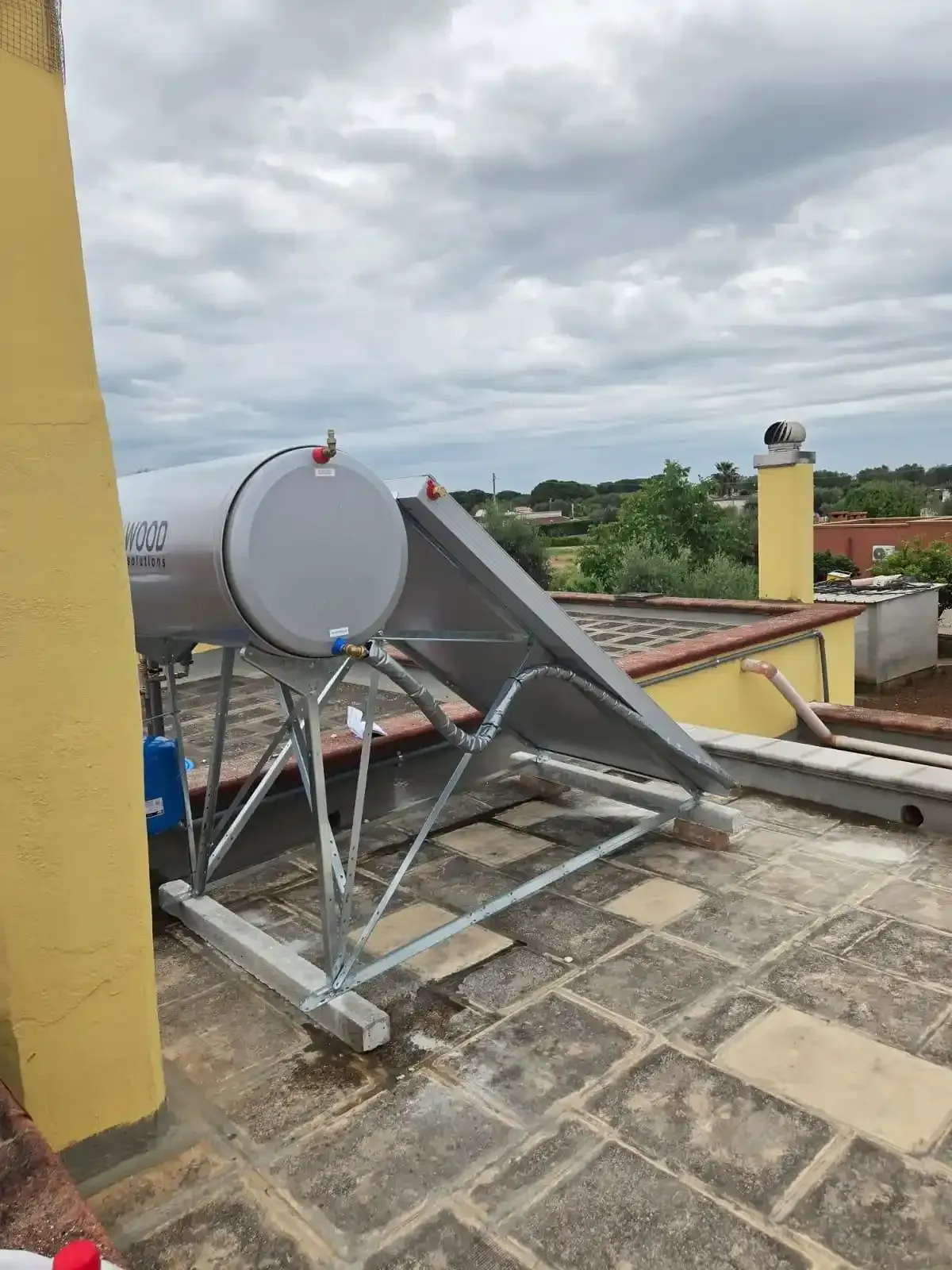 Scaldabagno solare da tetto con serbatoio e pannelli argentati su una terrazza piastrellata sotto un cielo nuvoloso.