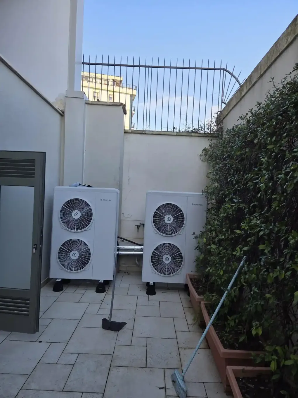 Due unità di climatizzazione esterne su uno stretto patio piastrellato, accanto a un muro bianco e una siepe.