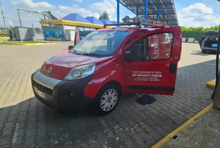 Un furgone rosso parcheggiato su un piazzale asfaltato con la portiera anteriore lato passeggero aperta, vicino a una stazione di servizio.