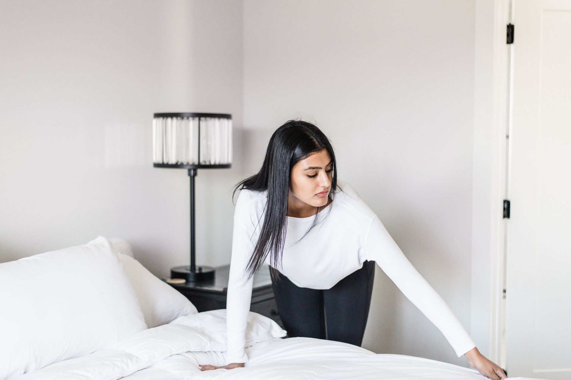 Woman in a white shirt and dark pants making a bed in a neutral-colored room.