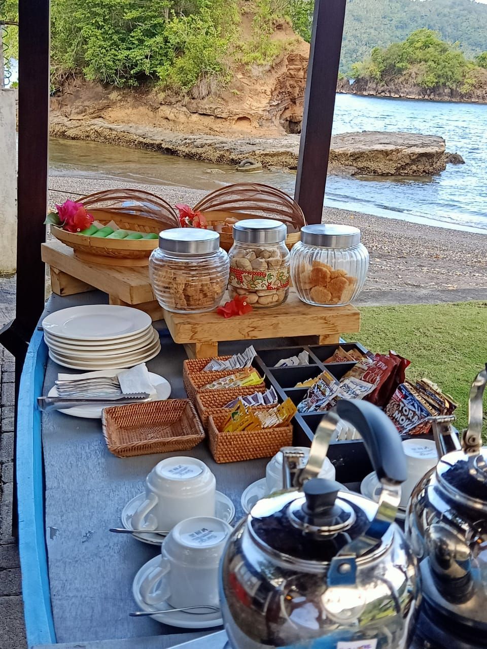 Outdoor breakfast buffet with pastries, plates, jars, and tea set on a table by the water