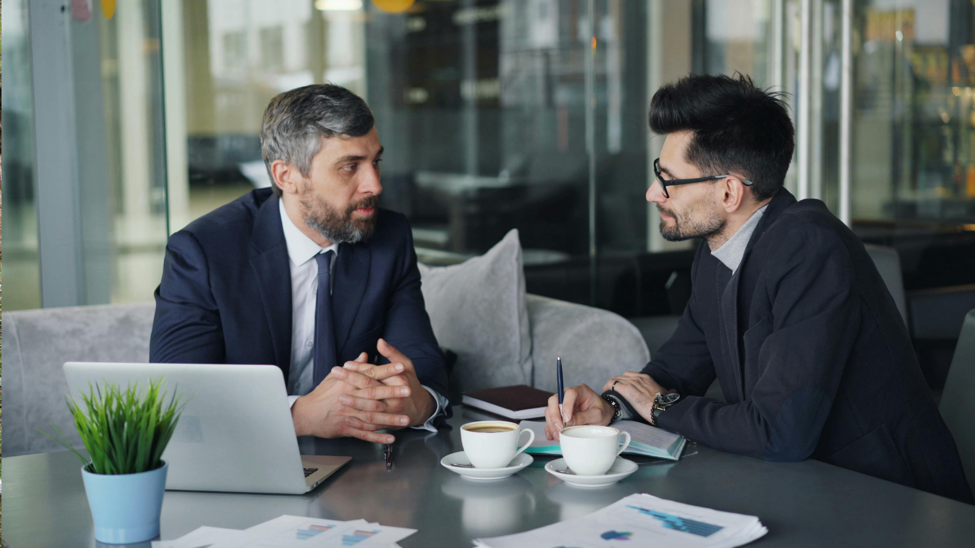Two men in suits engaged in a discussion at a table with coffee cups and a laptop in a cafe setting.
