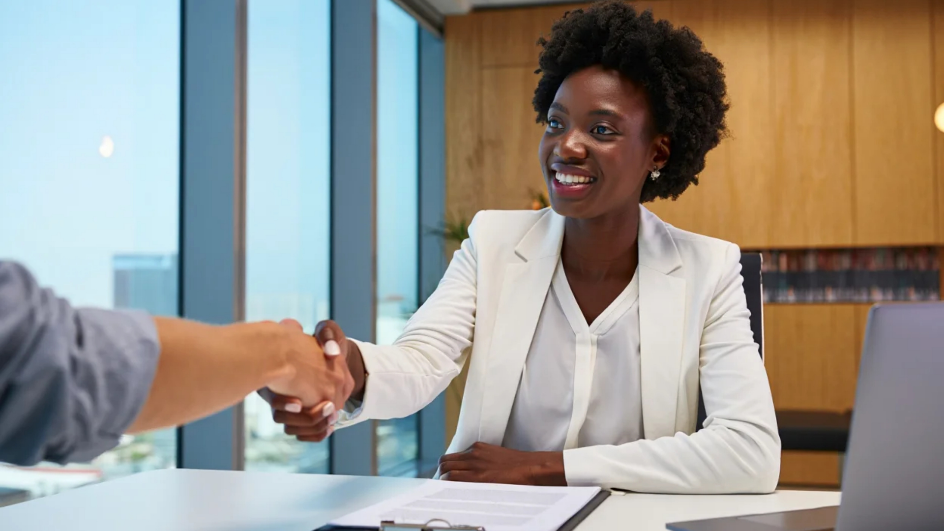 Woman in white suit shaking hands across a desk in an office setting.
