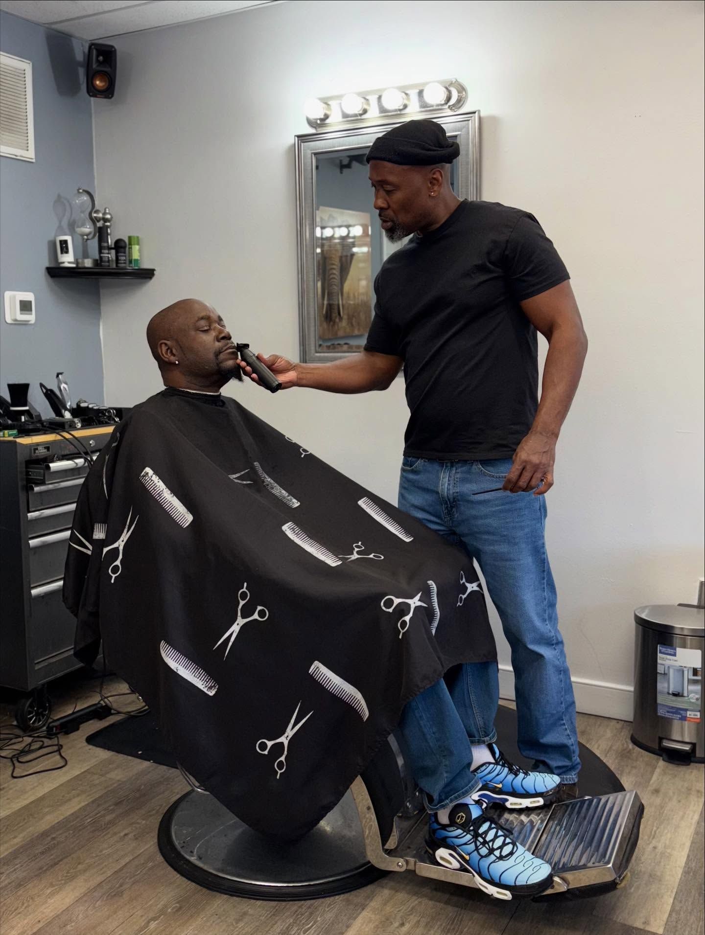 Barber trimming a customer’s beard in a barbershop, with the client seated under a black cape.