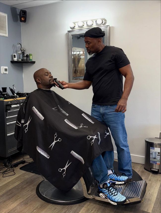 Barber trimming a customer’s beard in a barbershop, with the client seated under a black cape.