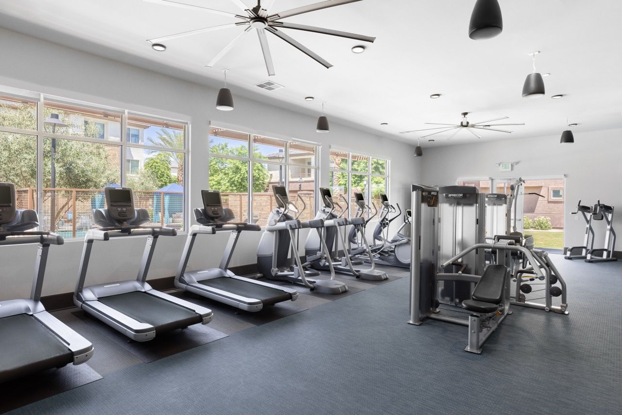 Bright community gym with a row of treadmills and cardio machines by large windows.