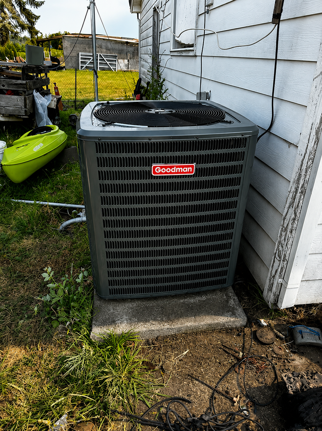 Outdoor air conditioner unit beside a white house wall on a concrete pad, with yard equipment nearby