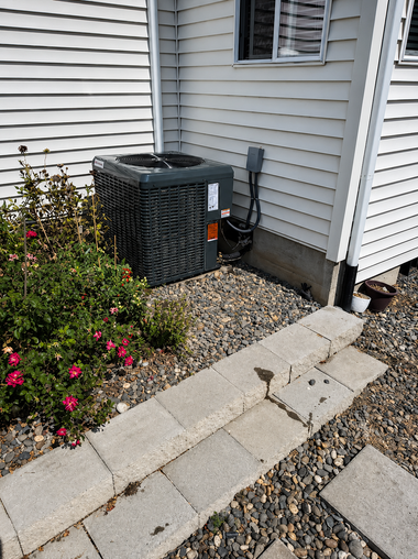 Outdoor air conditioner unit beside a house wall, next to gravel and stone pavers.