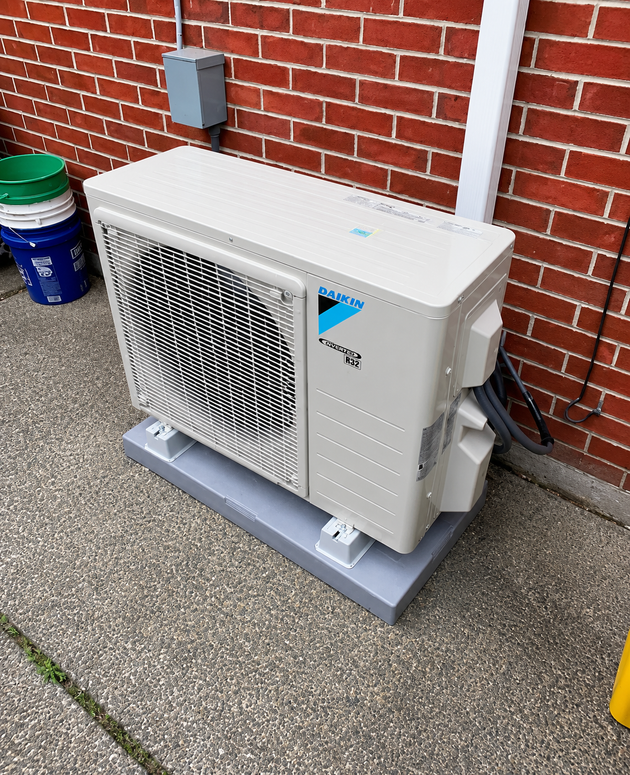 Outdoor HVAC unit beside a brick wall, mounted on a gray platform near a blue bucket.