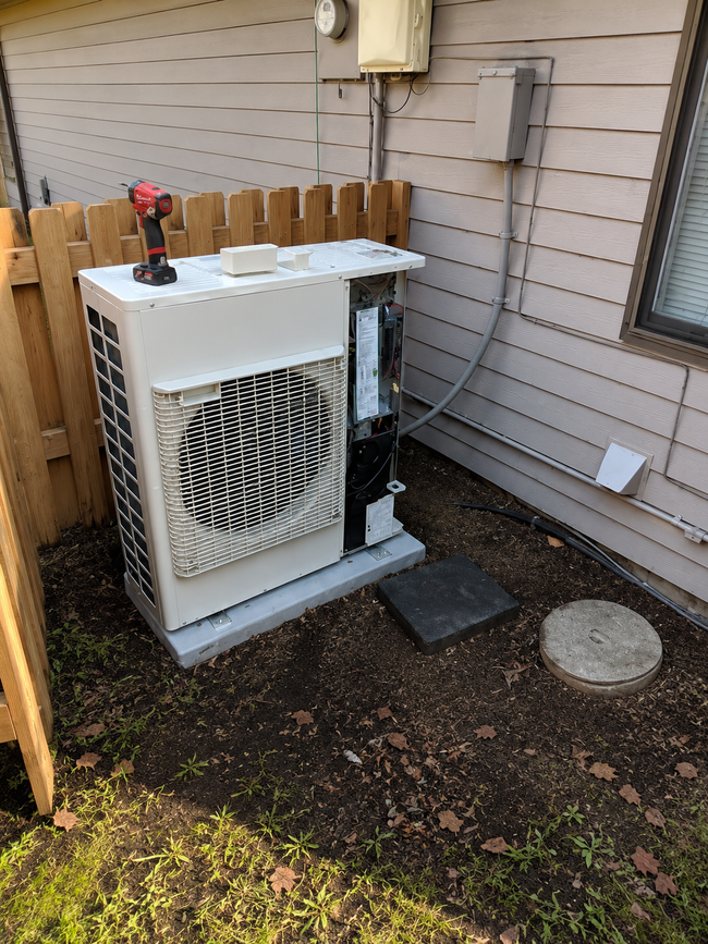 Outdoor HVAC unit beside a house wall, with tools and items stored on top.