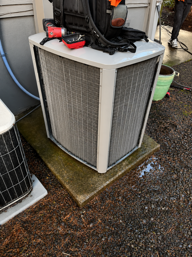 Outdoor HVAC condenser unit with a white grille on a concrete pad, wet ground around it