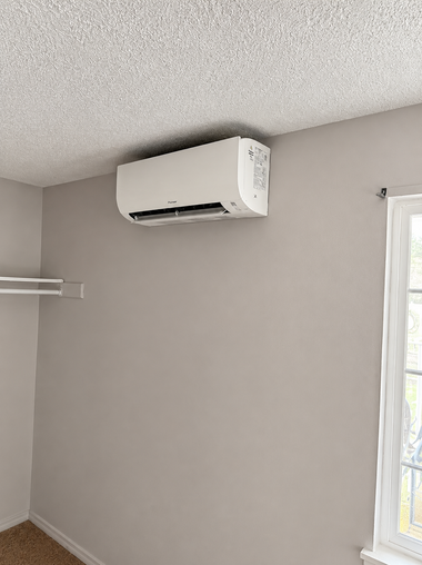 Wall-mounted white air conditioner on a beige wall beneath a textured ceiling, beside a window