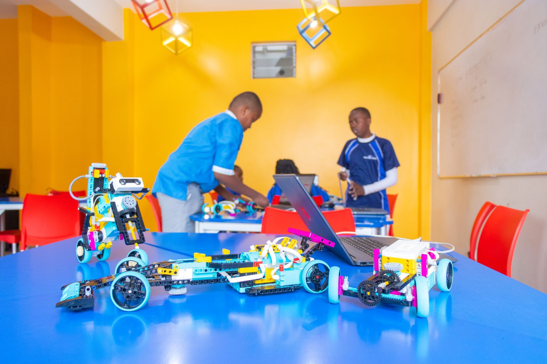 Students building robotics kits in a bright yellow room with blue tables, laptops, and unfinished mechanical models.
