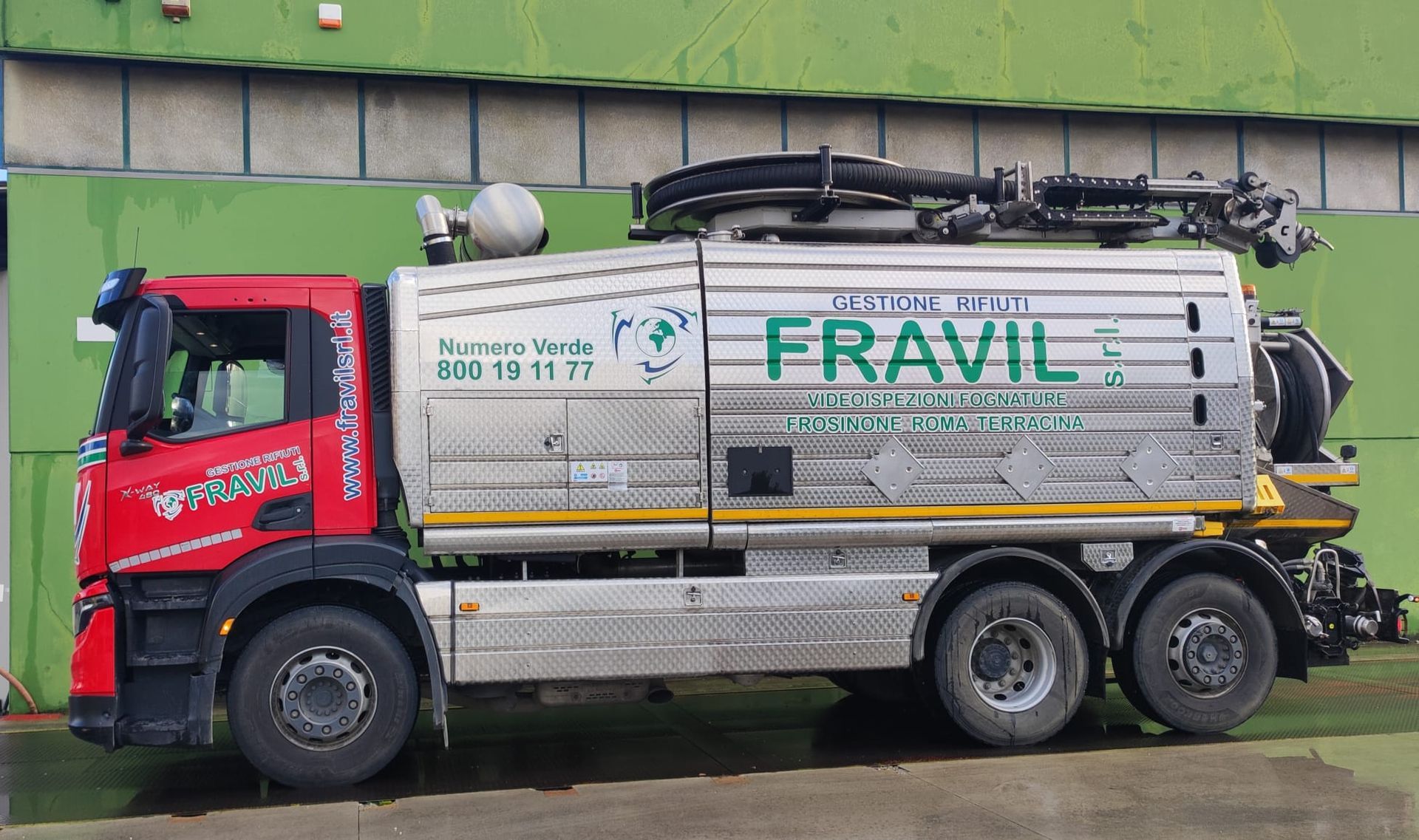 Camion FRAVIL rosso e argento con tubi flessibili, per la pulizia industriale, parcheggiato davanti a un muro verde.