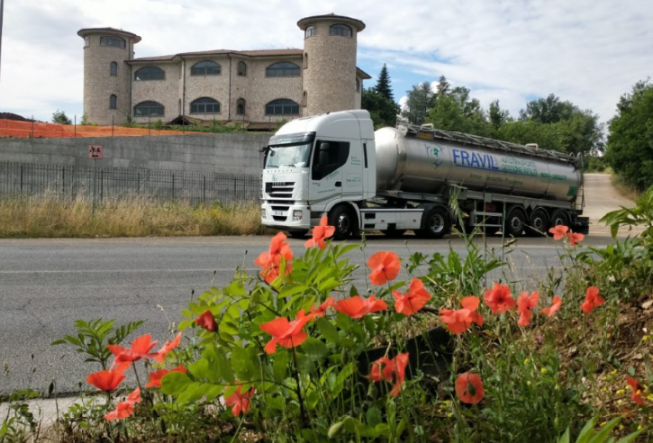 Autocisterna bianca FRAVIL sulla strada, papaveri in primo piano, grande edificio sullo sfondo.