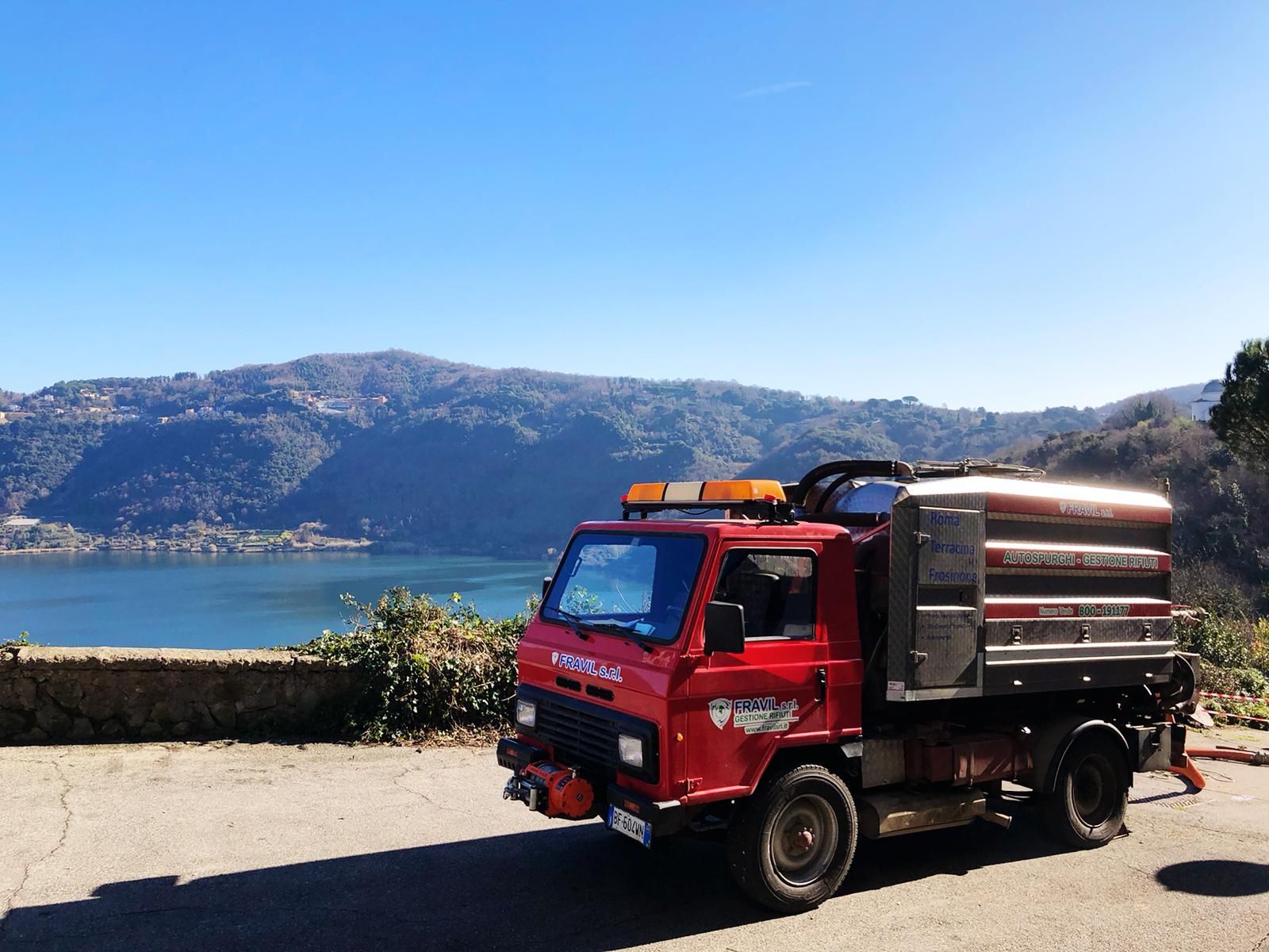 Un camion Fravil rosso parcheggiato su una strada che domina un lago e delle colline sotto un cielo azzurro.