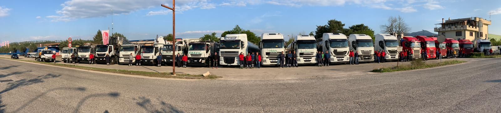 Una lunga fila di camion parcheggiati su un piazzale ghiaioso sotto un cielo parzialmente nuvoloso.
