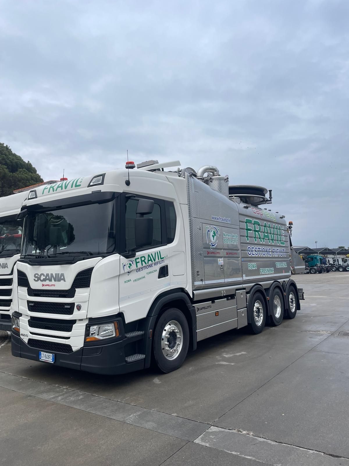Autospurgo Scania bianco con logo aziendale verde, parcheggiato su una superficie di cemento grigio. Cielo coperto.