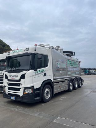 Autospurgo Scania bianco con logo aziendale verde, parcheggiato sul cemento sotto un cielo nuvoloso.