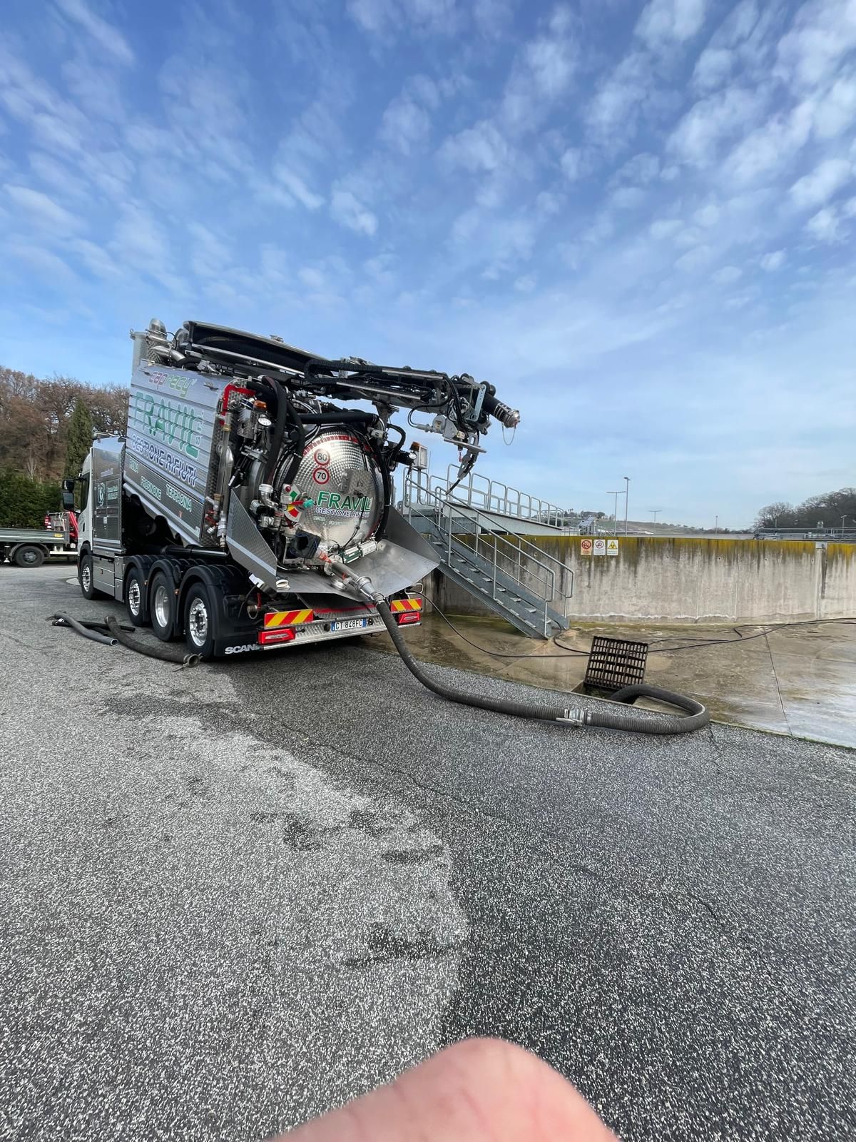 Camion settico, grigio scuro, che pompa liquido da una struttura in cemento sull'asfalto. Cielo azzurro.