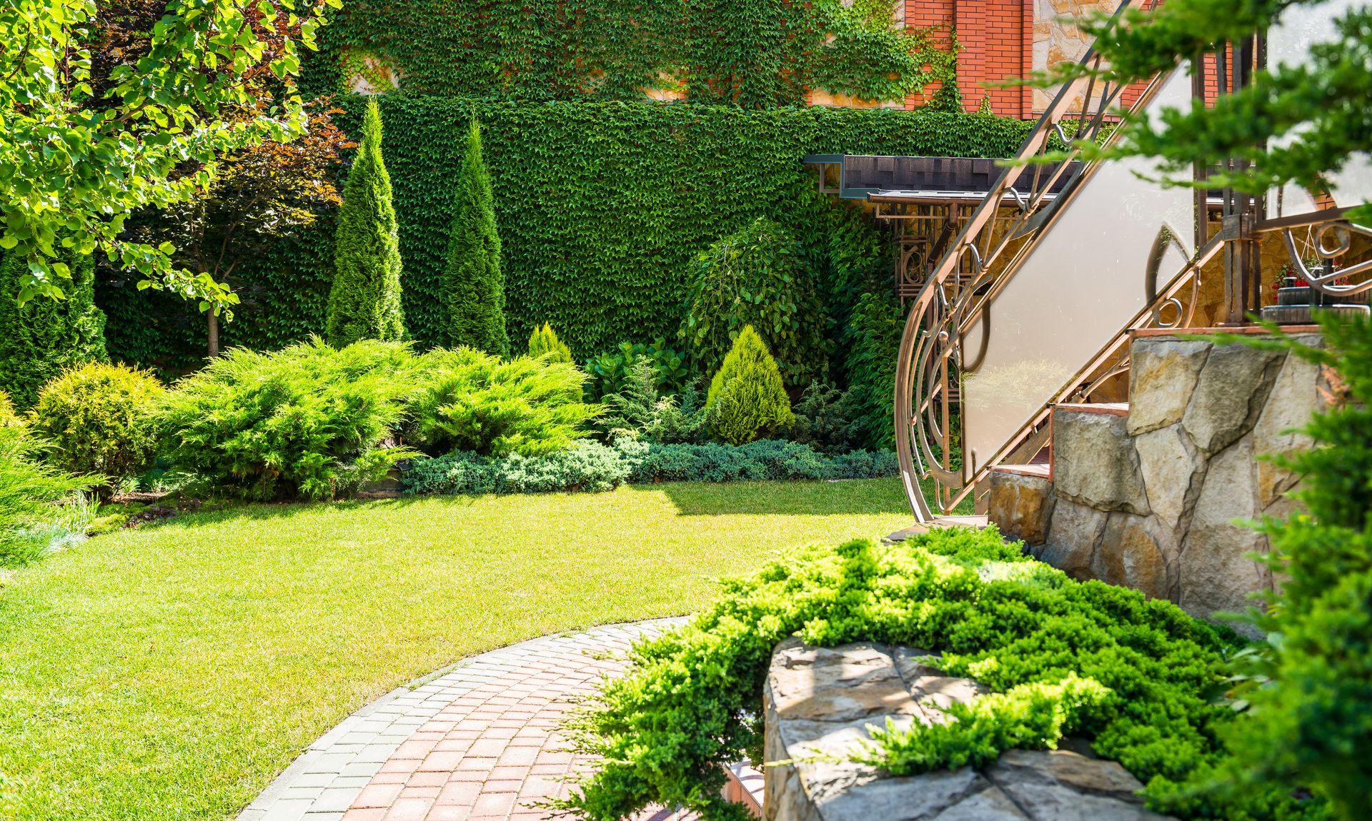 Lush garden with green lawn, ivy-covered wall, curved stone path, and stairs beside a stone retaining wall.