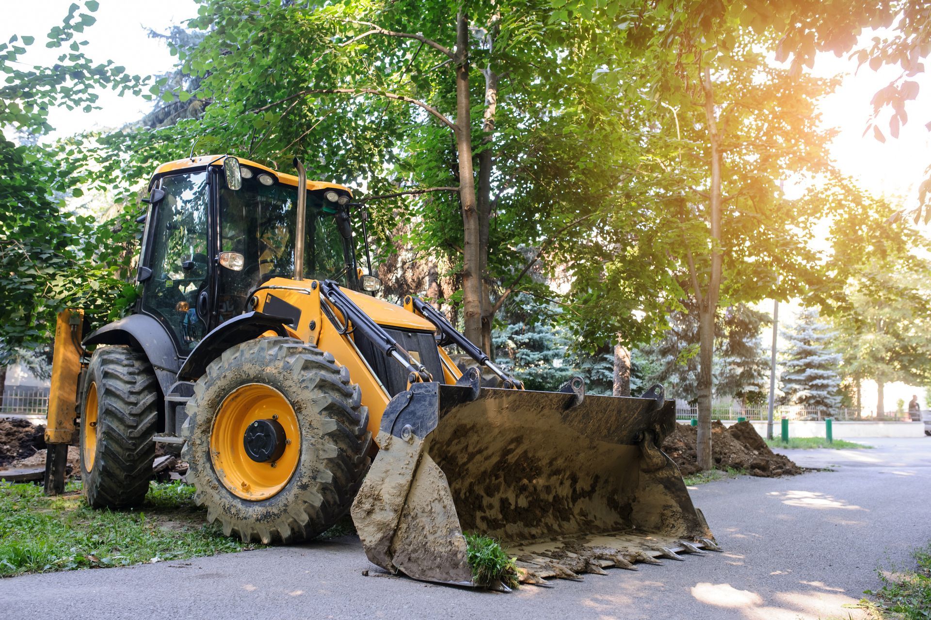 Yellow excavator lifting dirt on a tree-lined street in bright sunlight