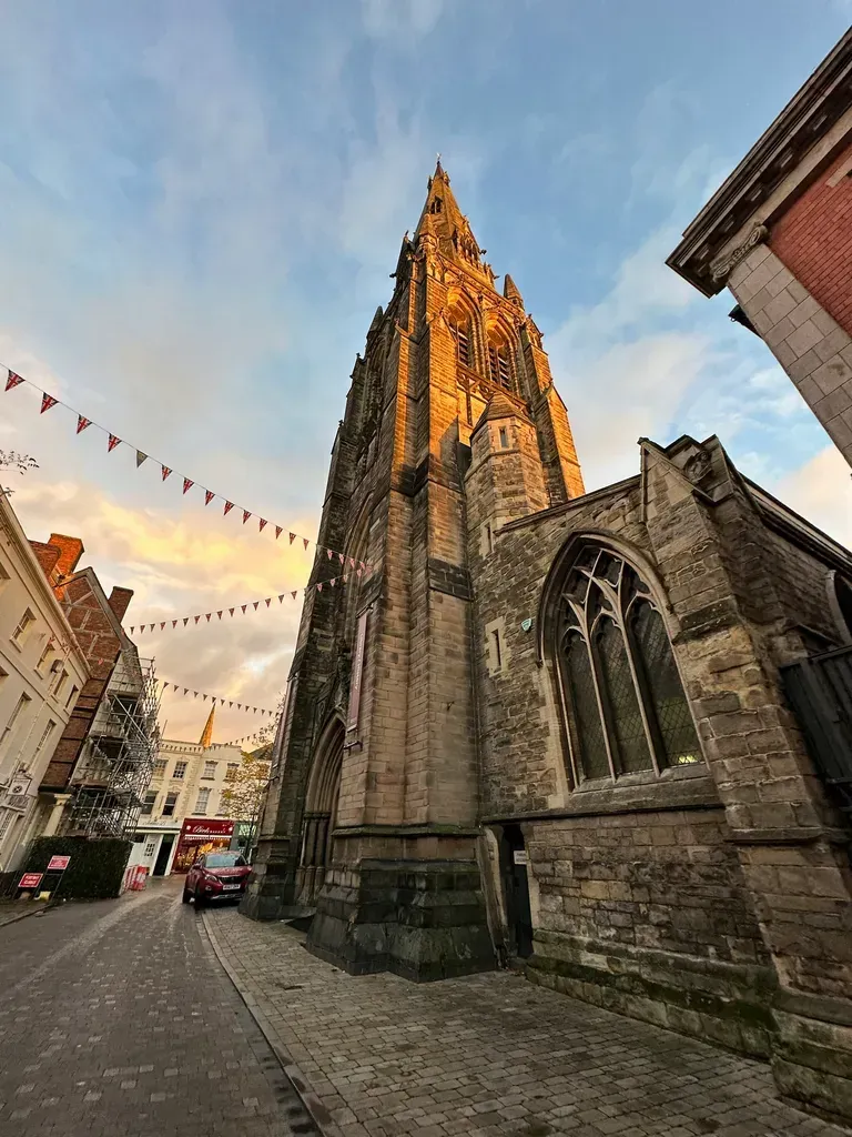 Gothic stone church tower on a narrow street at sunset, with bunting overhead and city buildings nearby