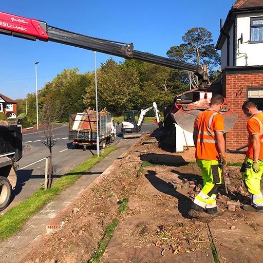 Roadside construction crew in orange gear working beneath a truck crane beside a house and excavator.
