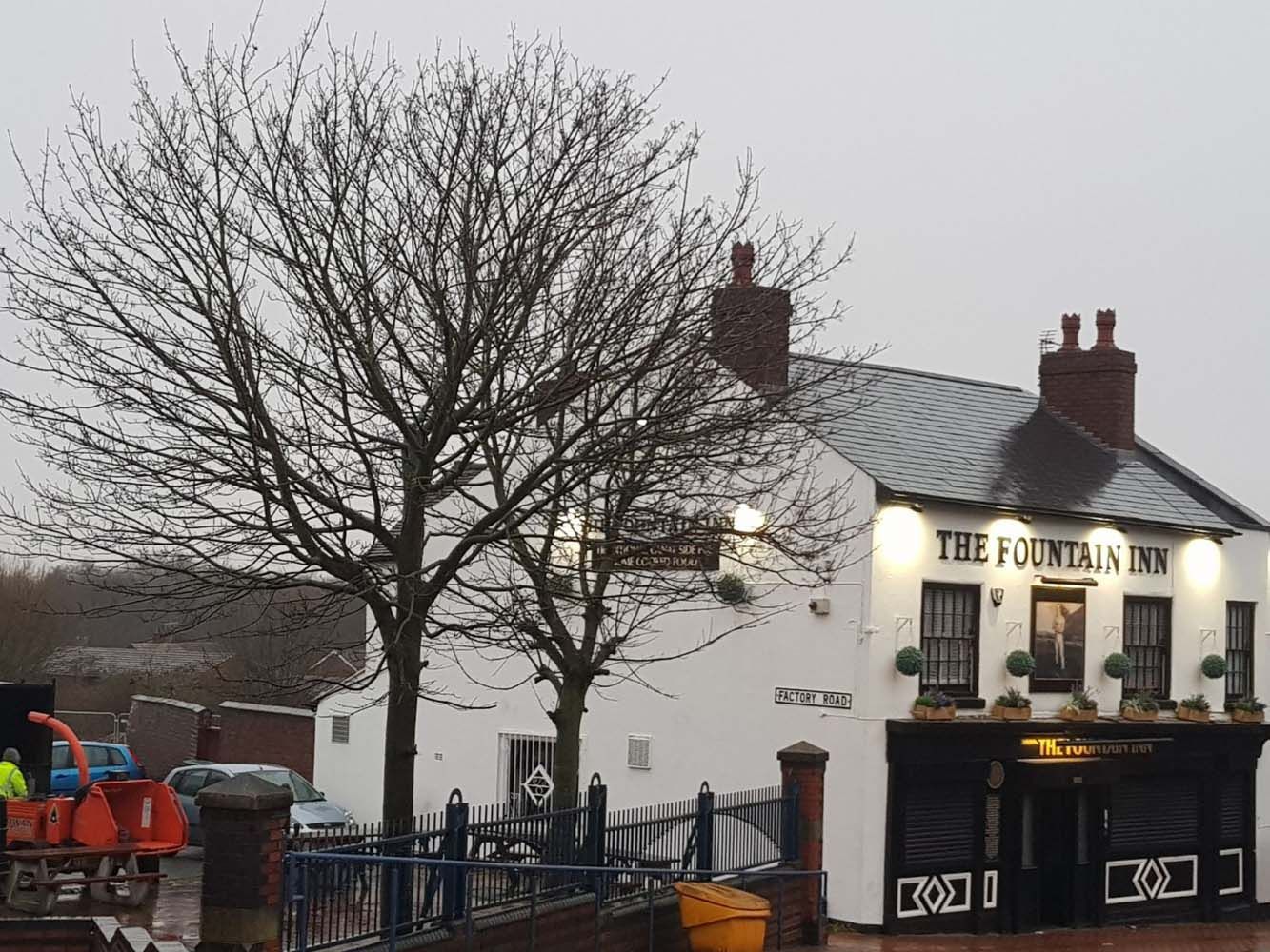 Small white pub with lit signs and a leafless tree on a rainy street corner.