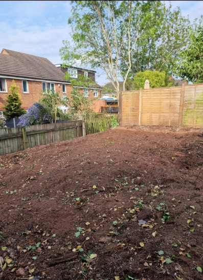 Freshly cleared garden bed with bare soil, wooden fence, brick houses, and trees in the background