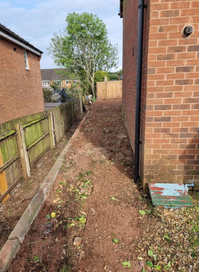Narrow dirt path between brick houses with a fence, leading to a tree and wooden gate.